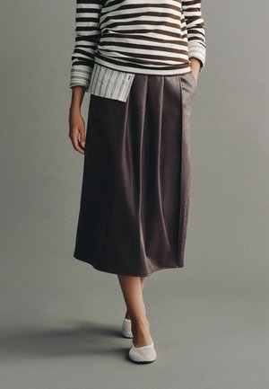 Person iført en brun midi nederdel, hvide mesh slip-on sko og en langærmet stribet top med én ærme rullet op.