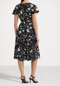 Černé květinové šaty s krátkými řasenými rukávy, přiléhavým korzetem a rozšířenou sukní. Celoplošný vzor bílých růží. Viditelné lodičky na vysokém podpatku.