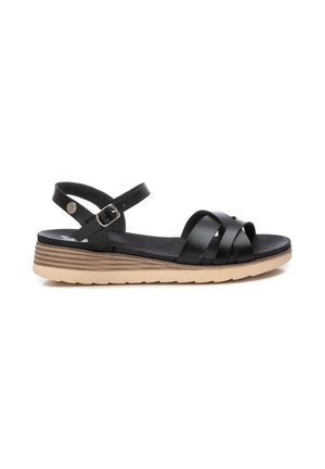 Sandalia plana de cuero negro con correas cruzadas, hebilla ajustable en el tobillo y suela texturizada de color marrón claro sobre fondo blanco.