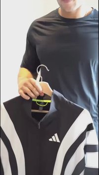 Mustvalge sportlik jakk, millel on tõmblukk ja kõrge krae, diagonaalsed triibud ning Adidas logo esiküljel, ripub naelal.