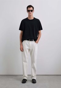 Tricou negru din bumbac, pantaloni albi din bumbac, centură neagră și pantofi negri. Modelul poartă ochelari de soare dreptunghiulari negri, stând în fața unui fundal simplu.