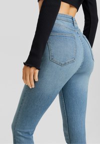 Pantalones vaqueros de tiro alto en azul claro con un corte ajustado, que cuentan con dos bolsillos traseros y una textura de denim suave. El diseño incluye una cinturilla visible.