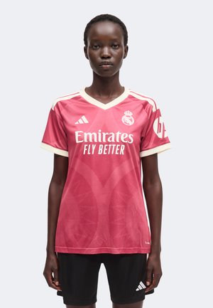 Mujer de pie con una camiseta rosa del Real Madrid con "Emirates Fly Better" y pantalones cortos negros de Adidas sobre un fondo liso.