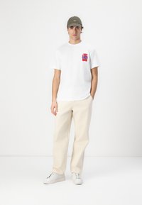 White short-sleeve T-shirt with a red and blue graphic on the left chest, paired with beige pants and white sneakers.