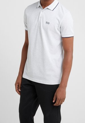 Homme portant un polo gris clair avec garniture noire et logo noir Hugo Boss sur la poitrine, assorti à un pantalon noir.