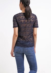 Femme avec une queue de cheval noire portant un haut en dentelle bleu marine à manches courtes et un jean bleu, de dos contre un fond blanc uni.