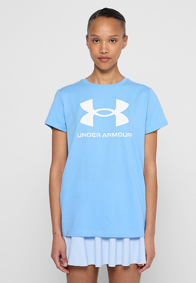 Młoda kobieta w jasnoniebieskiej koszulce Under Armour z białym logo i jasnoniebieskiej spódnicy, stojąca na jednolitym szarym tle.