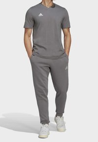 Chándal deportivo gris hecho de una tela suave. Parte superior de manga corta con cuello en V, pantalones ajustados a juego y zapatillas blancas. Logo de Adidas visible.