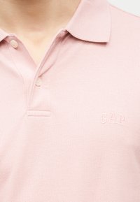 Polo rose clair avec deux boutons ouverts et logo embossé "GAP" sur le côté gauche de la poitrine.