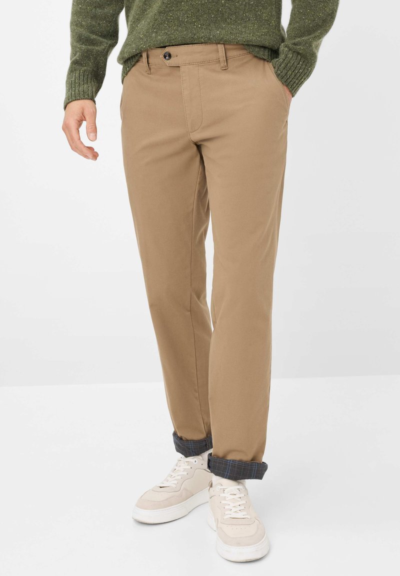Khakifarbene Chinos mit geradem Bein, ausgestattet mit einem Knopfverschluss, seitlichen Taschen und hochgekrempelten Säumen über weißen Sneakers.