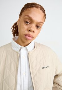 Giubbotto bomber trapuntato beige con zip frontale, dotato di un logo nero. Sotto, è visibile una camicia bianca a righe con bottoni.