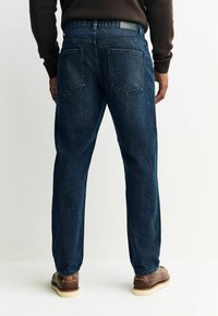 Mees, kes kannab tumedast helesinisest lõdvasti sobivaid teksapükse ja pruune nahkkingi, seisab lihtsa heleda tausta ees, seljaga.