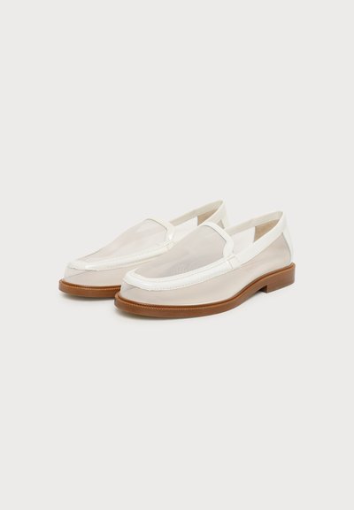 Paire de mocassins slip-on en mesh blanc avec semelles marron sur un fond uni clair, légèrement inclinée vers la gauche.