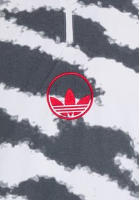 Grå och vit tie-dye pikétröja med dragkedja fram, som har en röd cirkulär Adidas-logotyp med tre blad på bröstet.