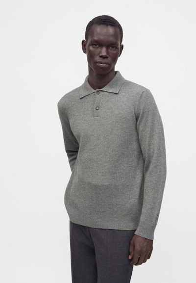 Homme à la peau foncée portant un polo en maille gris à manches longues et un pantalon sombre, debout devant un fond clair uni.