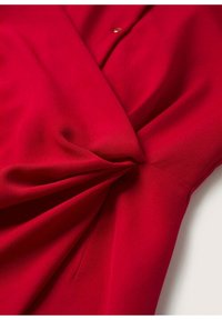 Blouse rouge en tissu lisse avec un détail torsadé à l'avant, dotée d'un accent bouton et d'un design drapé doux.