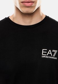 T-shirt nero a maniche corte con collo rotondo, con il logo bianco EA7 Emporio Armani sul lato sinistro del petto. Realizzato in materiale morbido e testurizzato.