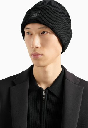 Beanie nero a coste con un'etichetta logo rettangolare. Indossato da un individuo vestito con una giacca nera e un top con zip.