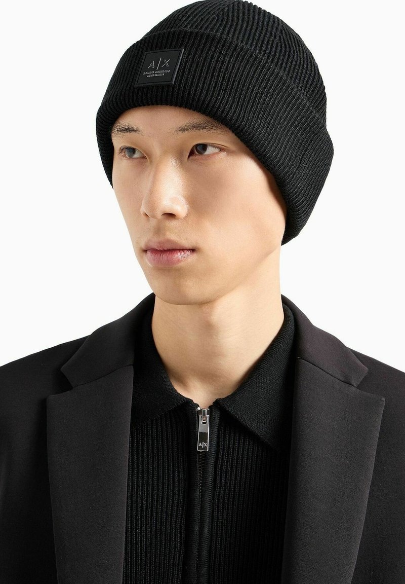 Beanie nero a coste con un'etichetta logo rettangolare. Indossato da un individuo vestito con una giacca nera e un top con zip.