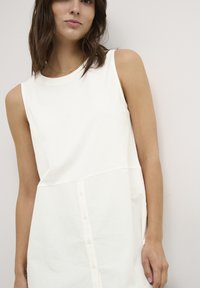 Un vestito bianco senza maniche con scollatura rotonda, caratterizzato da una chiusura frontale con bottoni e una vestibilità leggermente ampia, realizzato in un tessuto morbido e leggero.