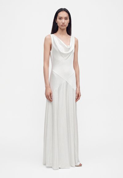 Woman with long dark hair wearing a sleeveless white satin gown with a draped neckline, standing against a plain light background.