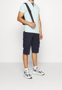 Chemise polo bleu clair avec un col marine, short cargo marine, et chaussures de sport multicolores, porté avec un sac à bandoulière noir.