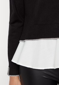 Suéter de punto negro con una capa de peplum blanca, con un ribete plateado en el dobladillo y en los puños. La tela parece suave y lisa.