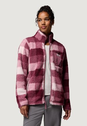 Mujer con chaqueta de forro polar de cuadros rosa y rojo oscuro sobre una camisa blanca y pantalones grises, de pie frente a un fondo liso.