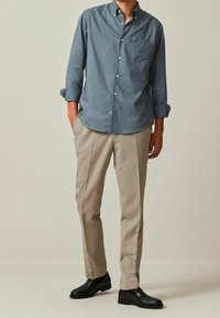 Chemise bleue boutonnée avec une poche poitrine, pantalon beige et chaussures de ville noires. La chemise a des manches retroussées et une texture lisse.