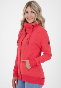 Woman with blonde hair smiling, wearing a bright coral zip-up jacket with high collar and blue jeans, hand in pocket.