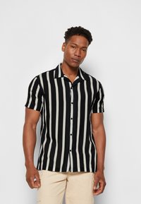 Chemise à manches courtes avec des rayures verticales noires et blanches, fermeture à boutons sur le devant et col de type camp. Fabriquée en tissu léger avec une texture lisse.