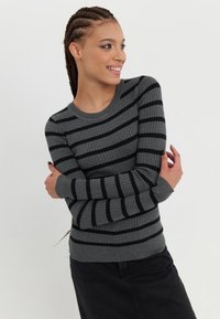Pull gris côtelé avec des rayures noires horizontales, manches longues et col rond. Modèle les bras croisés, portant une jupe noire.