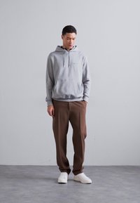 Sivi dukserica s prednjim kenguruvim džepom, uparena s smešnim pantalonama. Model nosi bele patike i stoji ispred neutralne pozadine.
