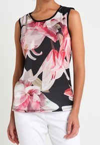 Débardeur noir sans manches avec un grand motif floral rose et blanc, porté avec un pantalon blanc, coupé au niveau du mi-torso.