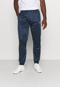 Pantaloni sportivi navy con una texture lucida e liscia, cintura elastica e polsini a coste. Indossati con scarpe sportive bianche con dettagli verdi.