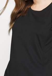 Donna che indossa una semplice maglietta nera attillata a maniche corte, con i lunghi capelli castani sulle spalle, su uno sfondo bianco.