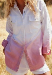 Femme portant une chemise à boutons blanche et rose dégradé avec les manches retroussées et un short blanc dans un environnement extérieur avec de l'herbe sèche.