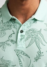 Vaaleanvihreä poolopaita, jossa on kuvioitu malli tummanvihrein kasvikuvioin. Kaksinappinen nappilista ja teksturoitu kangas.