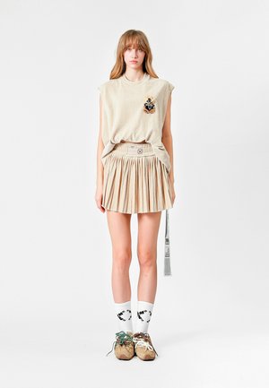 Top sin mangas de color beige con un parche decorativo, combinado con una falda plisada beige. La modelo lleva calcetines blancos y zapatillas de colores variados.