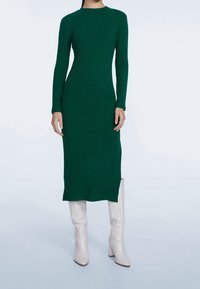 Robe en tricot côtelé vert avec des manches longues, présentant un col rond et une silhouette ajustée, associée à des bottes blanches à talons blocs montantes jusqu'aux genoux.