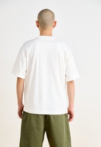 Camiseta blanca de algodón de manga corta con un ajuste relajado y cuello redondo, vista desde la espalda, combinada con pantalones verde oliva.