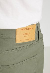 Pantalones de algodón verdes con un parche de cuero marrón. El parche presenta texto en relieve: "JACK & JONES, JEANS INTELLIGENCE, DESDE 1990."