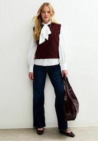 Burgundyröd stickad väst över en vit blus med slips, ihop med mörkblå utsvängda jeans och bruna ballerina-skor, hållande en burgundyröd tygväska.