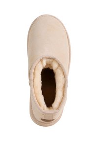 Beżowy zamszowy kapcie z miękką, futrzaną wyściółką i zaokrąglonym noskiem. Posiadają przeszywany szew wokół otworu i pętelkę na pięcie ułatwiającą zakładanie.