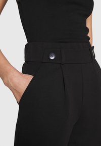 Detailní záběr osoby v černých kalhotách s vysokým pasem, které mají zapnutý pás, s rukou odpočívající v boční kapse, oblečené v černém topu bez rukávů.