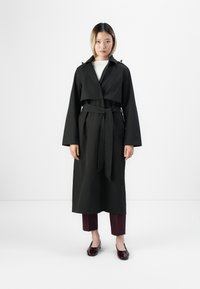 Donna in piedi con indosso un lungo trench coat nero con la vita legata, pantaloni corti bordeaux e mocassini lucidi bordeaux scuro.