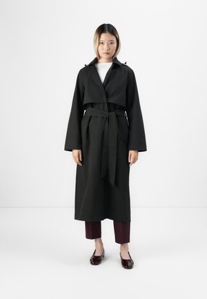 Donna in piedi con indosso un lungo trench coat nero con la vita legata, pantaloni corti bordeaux e mocassini lucidi bordeaux scuro.