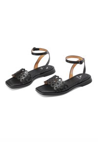 Sandali neri piatti presentano un design floreale decorativo intagliato sulla cinghia dell'alluce, cinghia alla caviglia regolabile e una suola rettangolare.