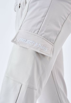 Pantaloni grigi chiari con una texture liscia. Presentano una tasca cargo sulla coscia con "PROJECT X PARIS" ricamato in bianco. Design snello.