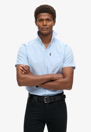 Camicia azzurro chiaro a maniche corte con colletto e taschino, abbinata a jeans neri e una cintura nera. Tessuto dalla trama liscia.